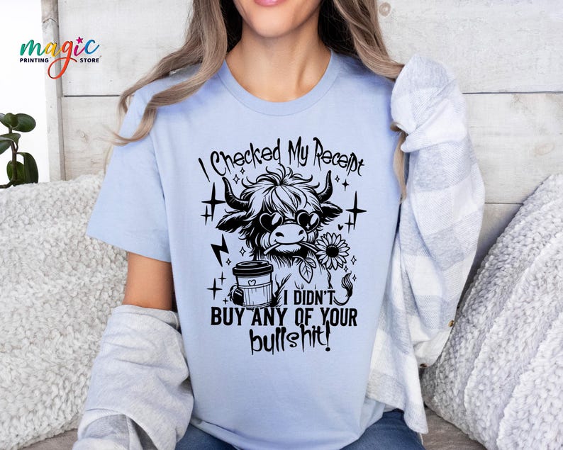 May include: Light blue t-shirt with a black and white graphic of a cow wearing sunglasses and holding a flower. Text on the shirt reads "I CHECKED MY RECEIPT I DIDN'T BUY ANY OF YOUR BULLSHIT!" The shirt has short sleeves.