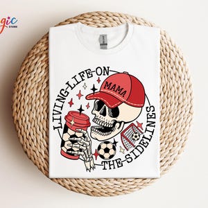 May include: White t-shirt featuring a graphic of a skull wearing a red baseball cap that says "Mama" and holding a coffee cup. The text around the skull reads "Living Life On The Sidelines".