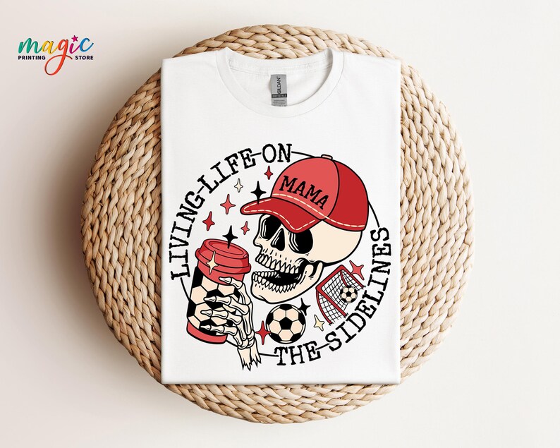 May include: White t-shirt featuring a graphic of a skull wearing a red baseball cap that says "Mama" and holding a coffee cup. The text around the skull reads "Living Life On The Sidelines".