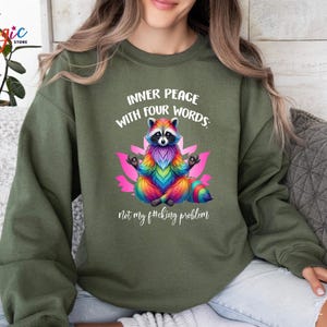 Pu&ograve; includere: Felpa verde oliva con un procione colorato in posa yoga, con la scritta "INNER PEACE WITH FOUR WORDS: not my f#cking problem". Il procione &egrave; raffigurato con pelliccia dai colori dell'arcobaleno ed &egrave; seduto su un fiore di loto rosa.