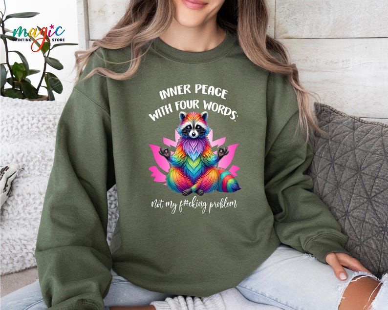 Pu&ograve; includere: Felpa verde oliva con un procione colorato in posa yoga, con la scritta "INNER PEACE WITH FOUR WORDS: not my f#cking problem". Il procione &egrave; raffigurato con pelliccia dai colori dell'arcobaleno ed &egrave; seduto su un fiore di loto rosa.
