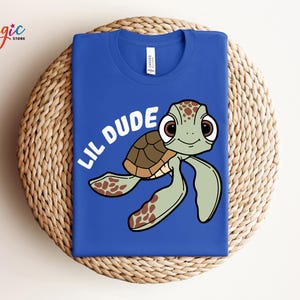 May include: Royal blue t-shirt featuring a cartoon turtle with the text "LIL DUDE" in white. The turtle has a brown shell, green flippers, and large brown eyes. The shirt is folded on a woven surface.