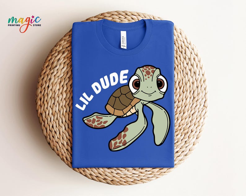 May include: Royal blue t-shirt featuring a cartoon turtle with the text "LIL DUDE" in white. The turtle has a brown shell, green flippers, and large brown eyes. The shirt is folded on a woven surface.