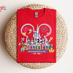 Puede incluir: Una camiseta roja con un gr&aacute;fico colorido de Disneyland. El dise&ntilde;o incluye el logotipo de Disneyland, un castillo, fuegos artificiales y varios personajes de Disney como Mickey Mouse, Donald Duck y Pluto. Tambi&eacute;n incluye el texto "The happiest place on earth".