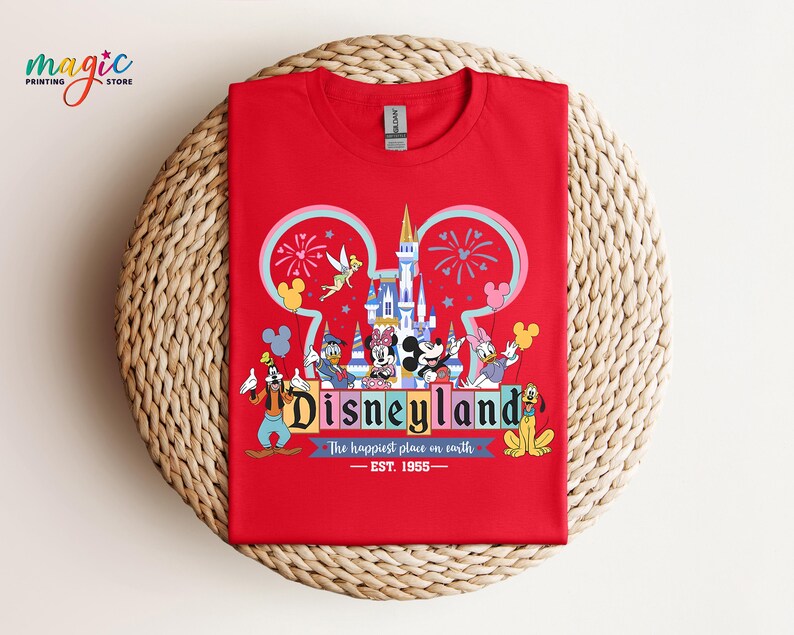 Puede incluir: Una camiseta roja con un gr&aacute;fico colorido de Disneyland. El dise&ntilde;o incluye el logotipo de Disneyland, un castillo, fuegos artificiales y varios personajes de Disney como Mickey Mouse, Donald Duck y Pluto. Tambi&eacute;n incluye el texto "The happiest place on earth".