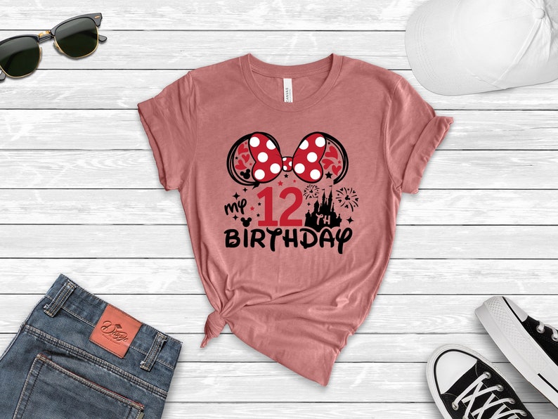 Pu&ograve; includere: Maglietta rosa con un fiocco rosso a pois bianchi e il testo "my 12th birthday" in nero. Il fiocco ha pois bianchi. Il testo include anche una piccola immagine della testa di Topolino e una silhouette del castello di Cenerentola.