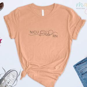 May include: A peach colored t-shirt with the text "NICU RN" and a line drawing of a baby in a heart shape.