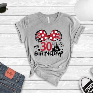 May include: Gray t-shirt with a red and white Minnie Mouse bow, a red number 30, and the text "my 30th birthday" in black. The design also includes a black silhouette of Cinderella's castle and fireworks.