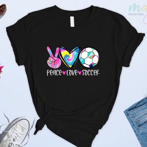 Może przedstawiać: Czarny T-shirt z symbolem pokoju, sercem i piłką nożną. Serce jest w stylu tie-dye, a tekst brzmi "Peace Love Soccer".