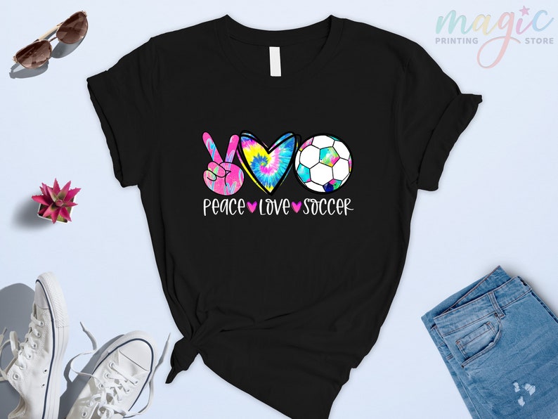 Może przedstawiać: Czarny T-shirt z symbolem pokoju, sercem i piłką nożną. Serce jest w stylu tie-dye, a tekst brzmi "Peace Love Soccer".