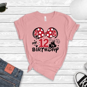 Pu&ograve; includere: Maglietta rosa con un fiocco di Minnie Mouse rosso e bianco, una grafica di castello nero e il testo "my 12th birthday".