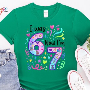 May include: A green t-shirt featuring the text "I was 67 Now I'm 67" in black. The numbers are adorned with colourful stars, hearts, and confetti. The shirt is styled with denim shorts and white trainers.