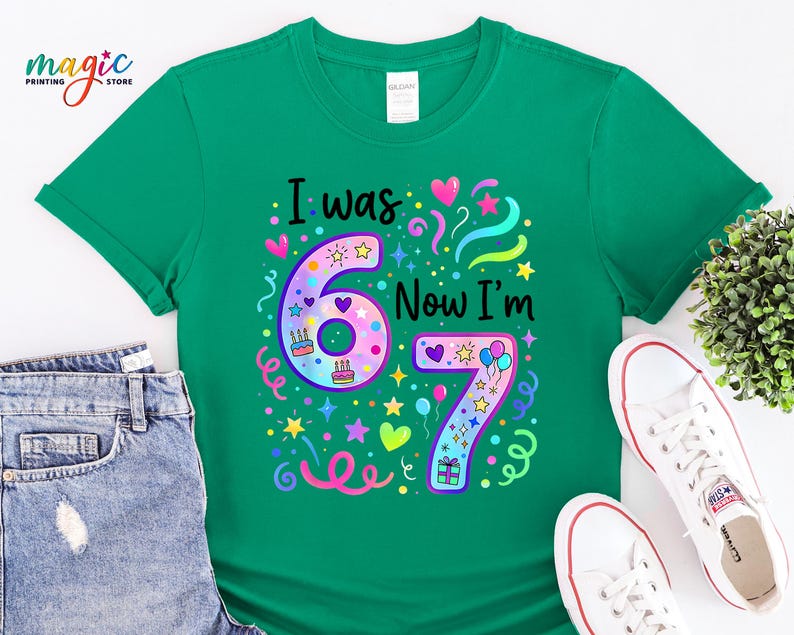 May include: A green t-shirt featuring the text "I was 67 Now I'm 67" in black. The numbers are adorned with colourful stars, hearts, and confetti. The shirt is styled with denim shorts and white trainers.
