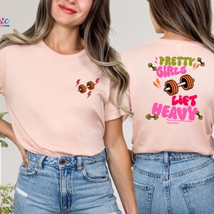 Puede incluir: Camiseta rosa claro con un peque&ntilde;o gr&aacute;fico de mancuernas y rayos en la parte delantera. La parte trasera de la camiseta presenta el texto "Pretty Girls Lift Heavy" con un gr&aacute;fico de mancuernas. La camiseta es de material suave.