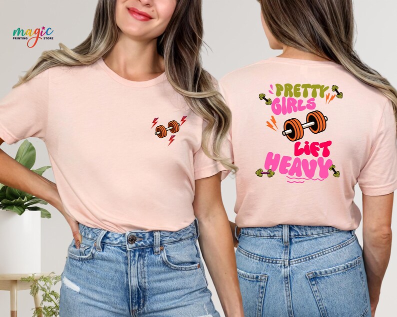 Puede incluir: Camiseta rosa claro con un peque&ntilde;o gr&aacute;fico de mancuernas y rayos en la parte delantera. La parte trasera de la camiseta presenta el texto "Pretty Girls Lift Heavy" con un gr&aacute;fico de mancuernas. La camiseta es de material suave.