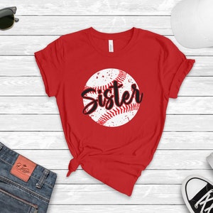 May include: Red t-shirt with a white baseball graphic and the word "Sister" in black lettering.