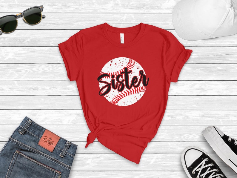 May include: Red t-shirt with a white baseball graphic and the word "Sister" in black lettering.