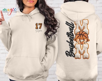 May include: Cream-colored hoodie with a front pocket and drawstring hood. The front features the number "17" in orange and black, with the name "Sophia" below. The back displays "Basketball Mom" with a basketball graphic.