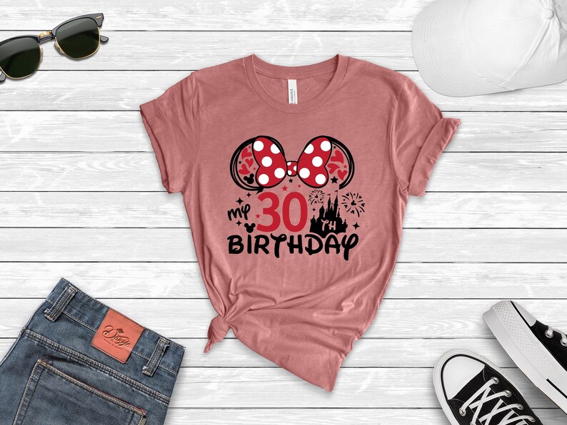 May include: A pink t-shirt with a black and white graphic of Minnie Mouse ears with a red bow and white polka dots. The graphic also includes the text "my 30th birthday" and a black silhouette of Cinderella's castle with fireworks.