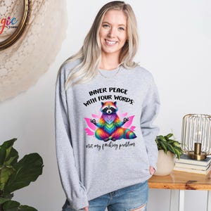 Pu&ograve; includere: Una felpa grigio chiaro con un procione colorato che medita su un fiore di loto rosa. Il testo "INNER PEACE WITH FOUR WORDS" e "Not my f*cking problem" &egrave; stampato sul davanti. Il procione &egrave; disegnato con colori arcobaleno.