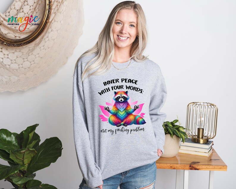 Pu&ograve; includere: Una felpa grigio chiaro con un procione colorato che medita su un fiore di loto rosa. Il testo "INNER PEACE WITH FOUR WORDS" e "Not my f*cking problem" &egrave; stampato sul davanti. Il procione &egrave; disegnato con colori arcobaleno.