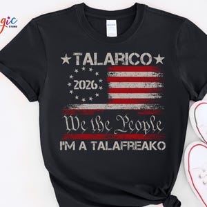 Può includere: Una t-shirt nera a maniche corte con la grafica di una bandiera americana invecchiata e il testo TALARICO, 2026, We the People e I'M A TALAFREAKO stampato in inchiostro bianco e rosso al centro del petto.
