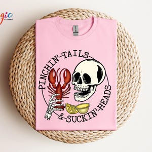 K&ouml;nnte beinhalten: Pinkes T-Shirt mit einem Grafikdruck eines Sch&auml;dels, der einen Hummer und Zitronen h&auml;lt. Der Text "Pinchin' Tails & Suckin' Heads" ist um die Grafik herum gedruckt.