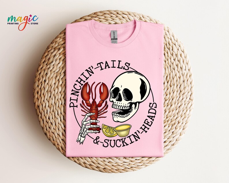 K&ouml;nnte beinhalten: Pinkes T-Shirt mit einem Grafikdruck eines Sch&auml;dels, der einen Hummer und Zitronen h&auml;lt. Der Text "Pinchin' Tails & Suckin' Heads" ist um die Grafik herum gedruckt.