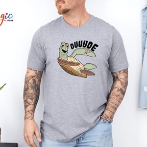 May include: A light gray t-shirt featuring a cartoon turtle with the word "DUUUUDE" in black. The turtle is green, brown, and tan. The shirt has short sleeves.