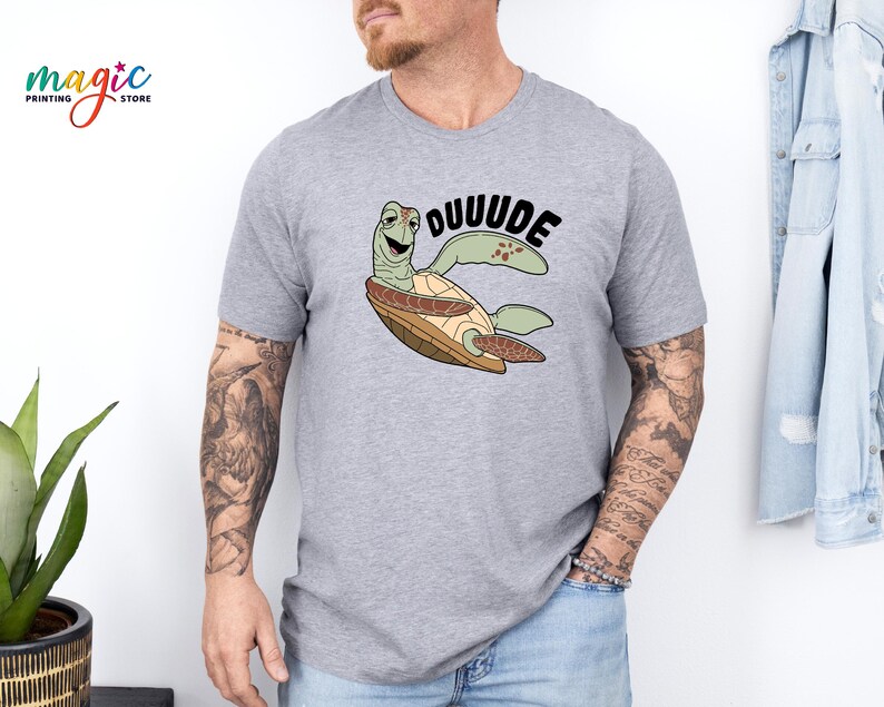 May include: A light gray t-shirt featuring a cartoon turtle with the word "DUUUUDE" in black. The turtle is green, brown, and tan. The shirt has short sleeves.