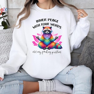 Pu&ograve; includere: Felpa bianca con una grafica colorata di procione in posa yoga, circondata da un fiore di loto rosa. Il testo recita: "INNER PEACE WITH FOUR WORDS: Not my f#cking problem."