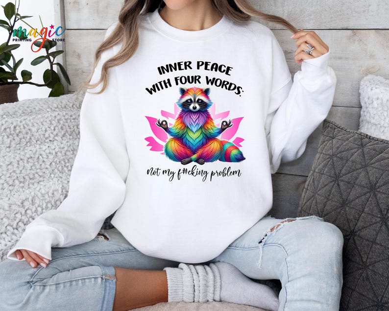 Pu&ograve; includere: Felpa bianca con una grafica colorata di procione in posa yoga, circondata da un fiore di loto rosa. Il testo recita: "INNER PEACE WITH FOUR WORDS: Not my f#cking problem."