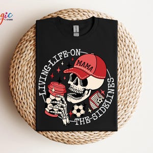 May include: Black t-shirt featuring a white skull wearing a red baseball cap with the word "Mama" on it. The skull is holding a coffee cup and a football. The text "Living Life On The Sidelines" is written in a circle around the skull.