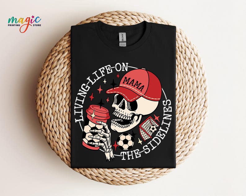 May include: Black t-shirt featuring a white skull wearing a red baseball cap with the word "Mama" on it. The skull is holding a coffee cup and a football. The text "Living Life On The Sidelines" is written in a circle around the skull.