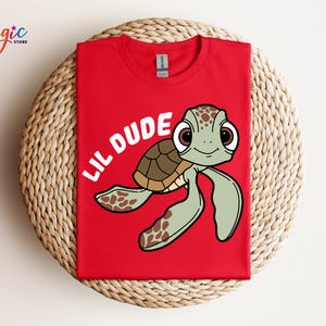 May include: Red t-shirt featuring a cartoon sea turtle with the text "LIL DUDE" in white. The turtle has a brown shell, green flippers, and large, expressive eyes. The shirt is made of a soft material.