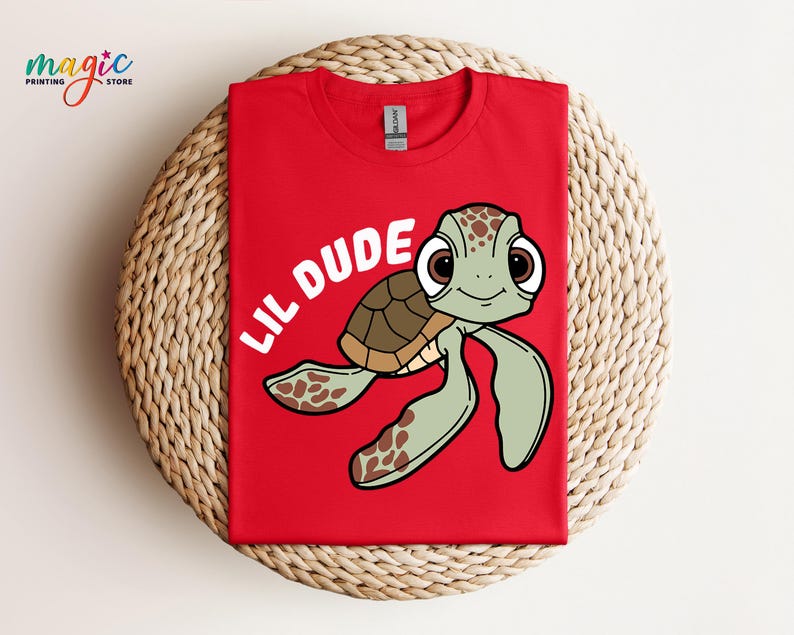May include: Red t-shirt featuring a cartoon sea turtle with the text "LIL DUDE" in white. The turtle has a brown shell, green flippers, and large, expressive eyes. The shirt is made of a soft material.