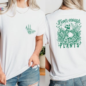 May include: White t-shirt with a green skeleton holding a watering can surrounded by plants and the text "Never enough plants".