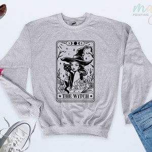 Può includere: Una felpa girocollo grigio melange con una stampa grafica nera raffigurante un design di carta dei tarocchi. L'illustrazione mostra una persona con un cappello da strega a tesa larga, che tiene in braccio un gatto nero, circondata da fiori, con il testo THE WITCH.