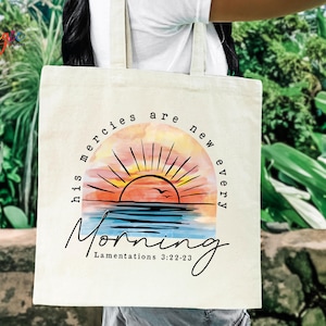 May include: Cream-colored tote bag featuring a watercolor design of a sunrise over the ocean. The text "his mercies are new every Morning" and "Lamentations 3:22-23" are printed in black script. The bag has two beige handles.