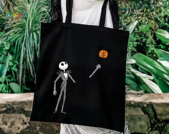 May include: A black tote bag featuring a white graphic of a tall, thin skeleton character in a pinstripe suit. The character holds a string attached to an orange pumpkin shaped like a mouse head. The bag has two black fabric shoulder straps.