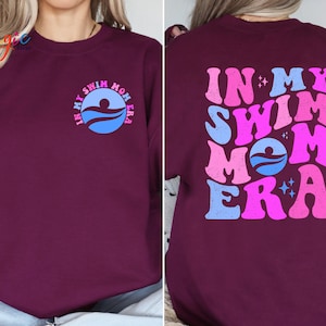 May include: Burgundy sweatshirt with the text "In My Swim Mom Era" in pink and blue lettering on the front and back. The back design features a stylised wave and swimmer graphic.