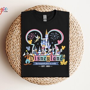 Puede incluir: Camiseta negra con un gr&aacute;fico colorido de Disneyland. El dise&ntilde;o incluye el logotipo de Disneyland, un castillo, fuegos artificiales y varios personajes de dibujos animados. Tambi&eacute;n se incluye el texto "The happiest place on earth - EST. 1955".