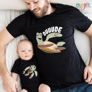 May include: Black t-shirt with a cartoon turtle and the word "OUUUDE" in white. A matching black baby onesie with a turtle graphic and the words "LIL DUDE". The shirts are likely for a parent and child.