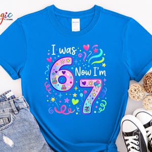 May include: A vibrant blue t-shirt featuring the phrase "I was 6 Now I'm 7" in a colourful design. The graphic includes the numbers 6 and 7, decorated with hearts, stars, and birthday cake illustrations. The shirt is paired with denim jeans and black trainers.