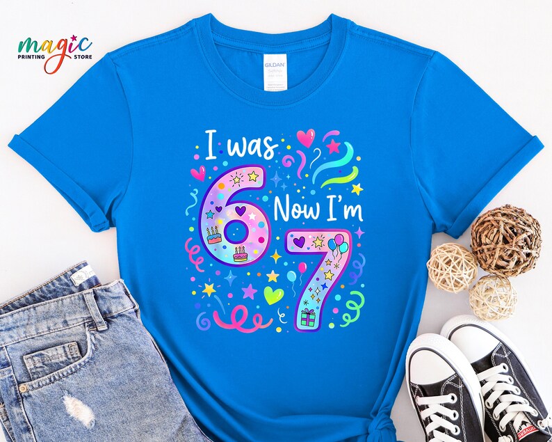 May include: A vibrant blue t-shirt featuring the phrase "I was 6 Now I'm 7" in a colourful design. The graphic includes the numbers 6 and 7, decorated with hearts, stars, and birthday cake illustrations. The shirt is paired with denim jeans and black trainers.