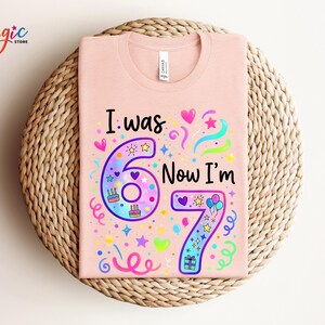 May include: A pale pink t-shirt featuring the phrase "I was 6 Now I'm 7" in a vibrant, child-like font. The design incorporates the numbers 6 and 7, adorned with hearts, stars, and cake illustrations. The t-shirt is presented on a woven mat.