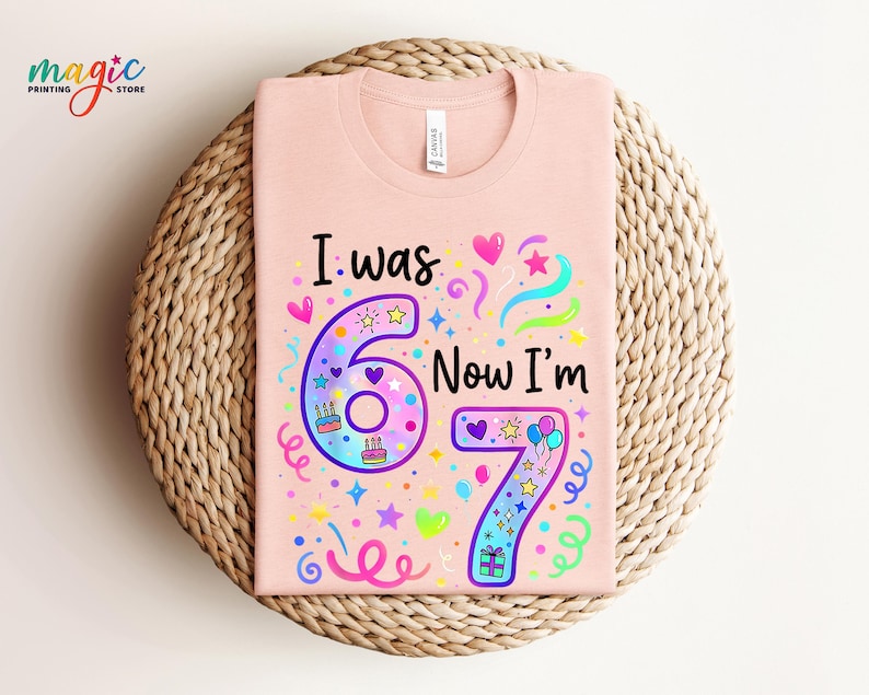May include: A pale pink t-shirt featuring the phrase "I was 6 Now I'm 7" in a vibrant, child-like font. The design incorporates the numbers 6 and 7, adorned with hearts, stars, and cake illustrations. The t-shirt is presented on a woven mat.