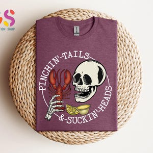K&ouml;nnte beinhalten: Ein maroonfarbenes T-Shirt mit einem wei&szlig;en Grafikdesign eines Sch&auml;dels, der einen Hummer mit dem Text "Pinchin' Tails & Suckin' Heads" h&auml;lt.