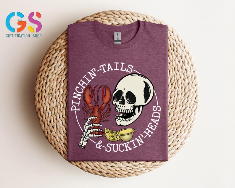 K&ouml;nnte beinhalten: Ein maroonfarbenes T-Shirt mit einem wei&szlig;en Grafikdesign eines Sch&auml;dels, der einen Hummer mit dem Text "Pinchin' Tails & Suckin' Heads" h&auml;lt.