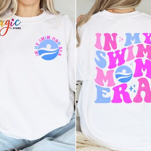 May include: White sweatshirt with a blue and pink graphic that says "In My Swim Mom Era" on the back. The front of the sweatshirt has a smaller blue graphic with a swimmer and the words "In My Swim Mom Era".